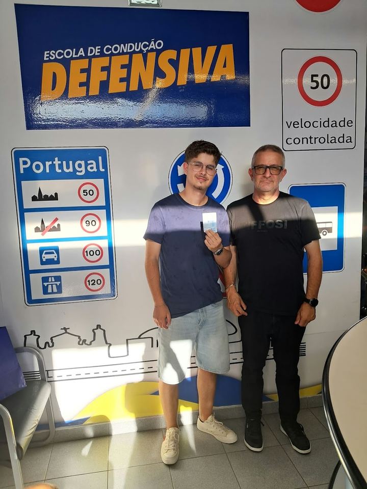 Novo condutor com o instrutor, a mostrar a carta em frente à parede de sinais da escola