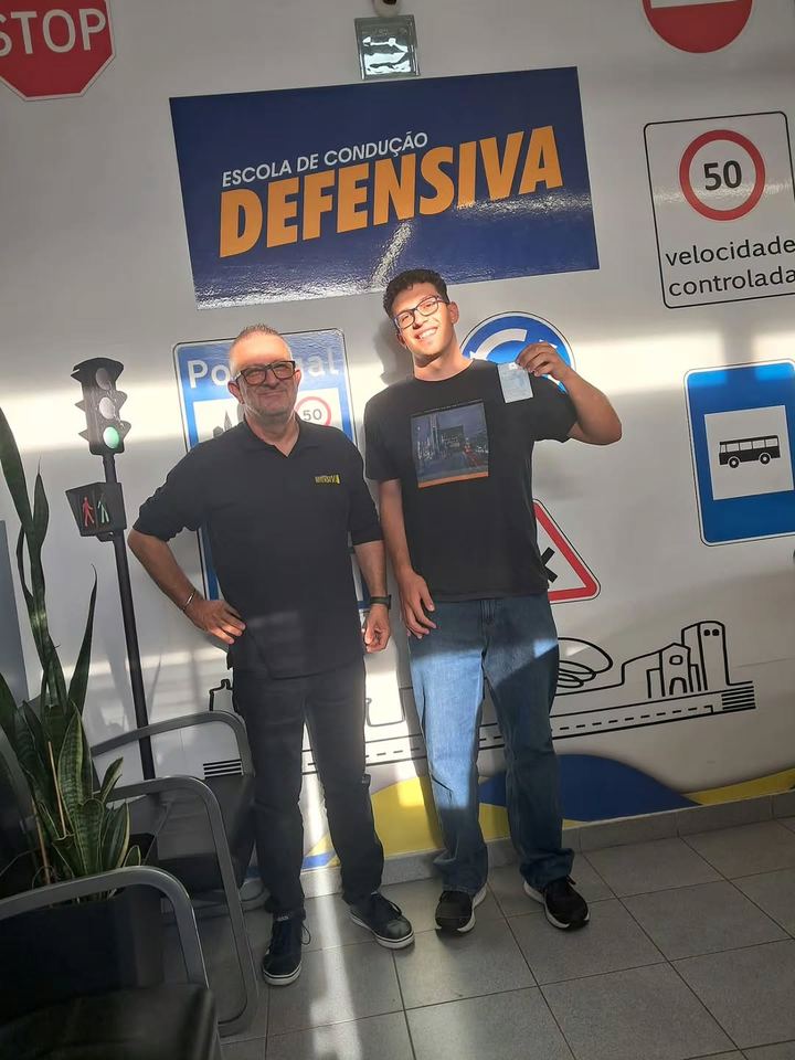 Novo condutor com o instrutor, a mostrar a carta em frente à parede de sinais da escola