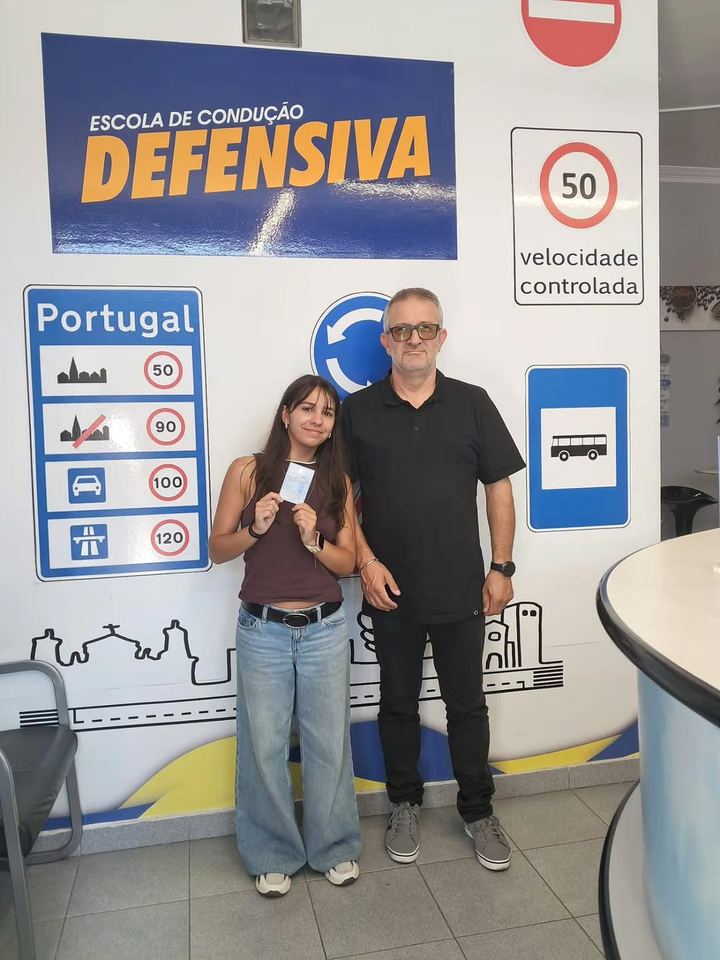 Nova condutora com o instrutor, a mostrar a carta em frente à parede de sinais da escola