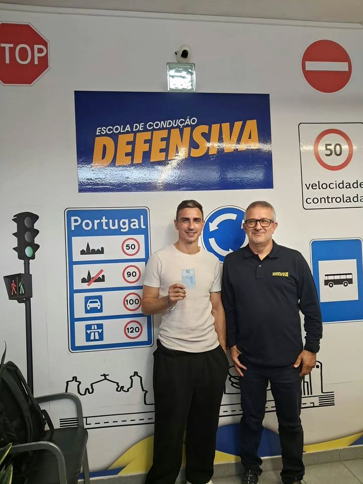 Novo condutor com o instrutor, a mostrar a carta em frente à parede de sinais da escola